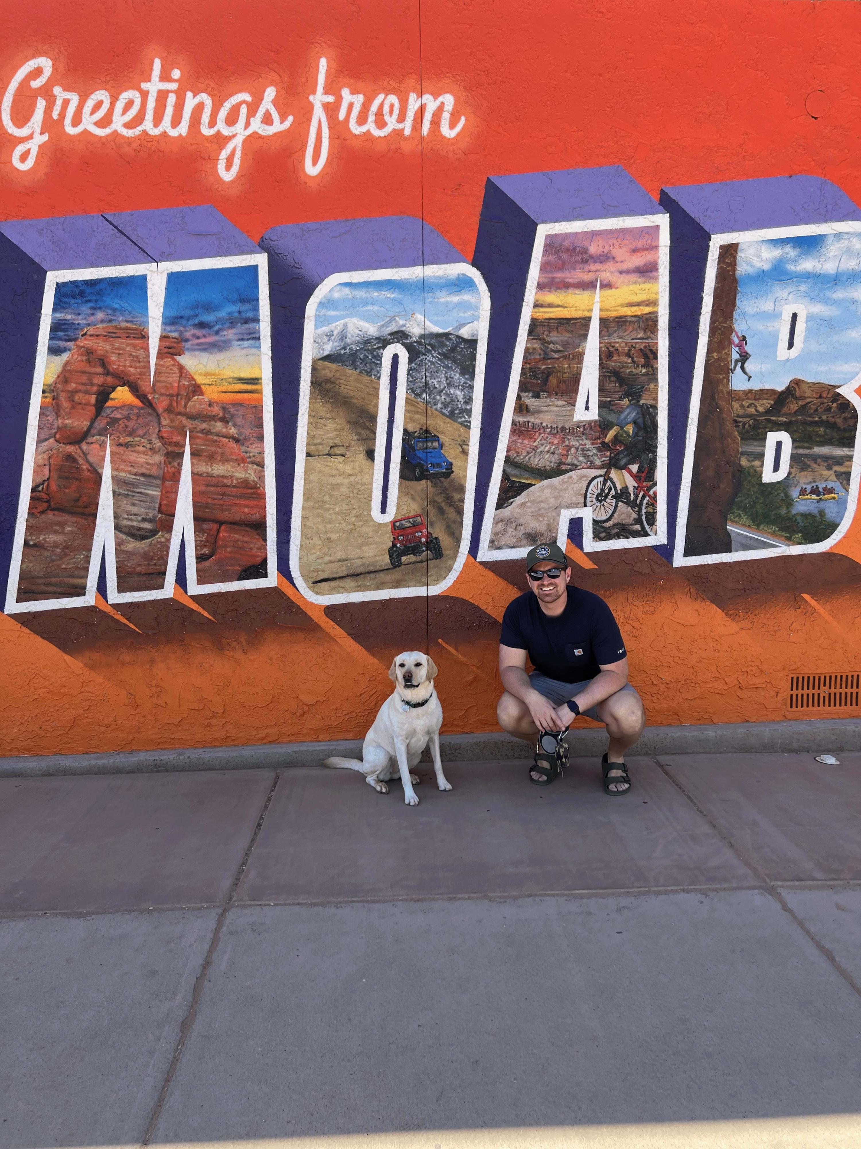 A man and a dog posing in front of a wall that reads "Greetings from MOAB"